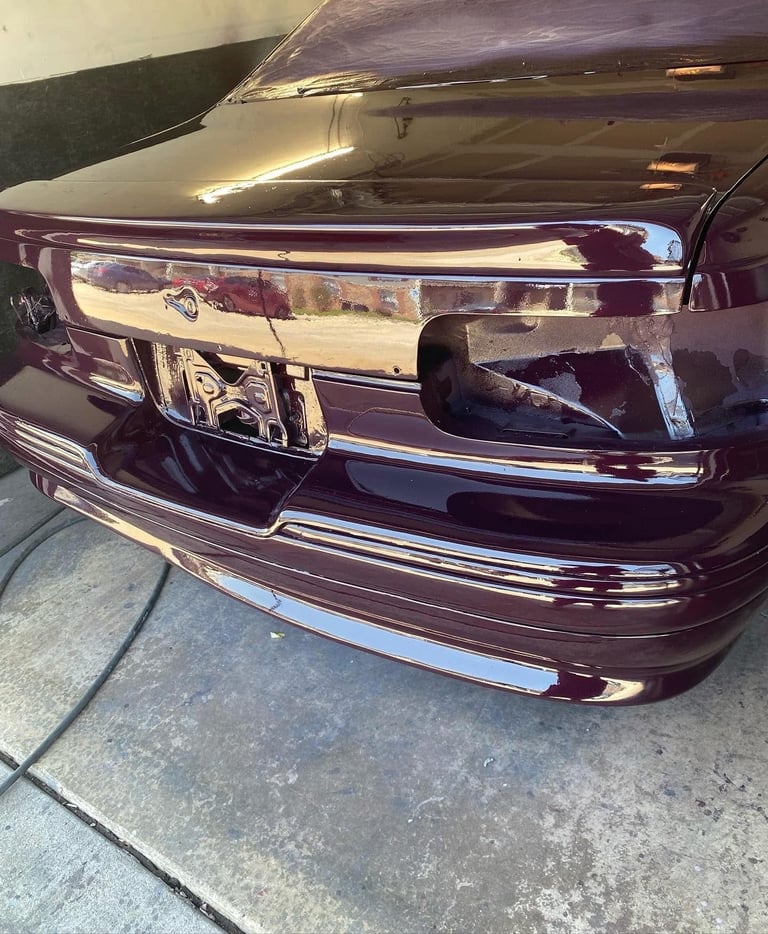 Close-up of a dark burgundy cars front bumper and headlight assembly with chrome trim on concrete pavement